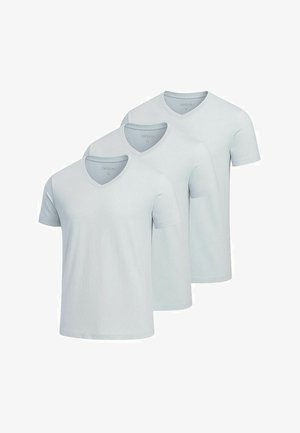Lyseblå V-hals t-shirt, mørk navy elastiske bukser, hvide slip-on sko; model står med armene krydset, minimal baggrund.