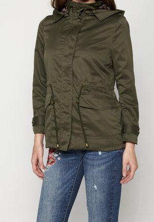 Žena nosící olivově zelenou bundu s kapucí, předními kapsami a stahovacími šňůrkami, kombinovanou s modrými džínami s květinovou výšivkou a otřepy.