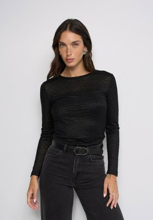 Mujer con cabello largo y castaño, vistiendo una blusa negra transparente de manga larga con textura, vaqueros negros de cintura alta, cinturón negro y pendientes plateados sobre un fondo blanco.