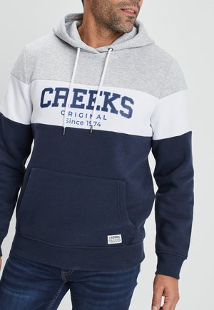 Felpa con cappuccio nei colori grigio, bianco e blu navy, con una tasca frontale, caratterizzata dal testo "CREEKS ORIGINAL Dal 1974" in blu. Materiale misto cotone.