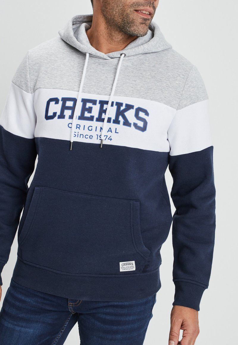 Kapuzensweatshirt in Grau, Weiß und Marineblau mit einer Vordertasche, versehen mit dem Text "CREEKS ORIGINAL Seit 1974" in Blau. Aus Baumwollmischung.