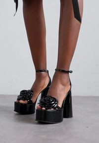 Sandales à talons compensés noires avec une finition brillante, présentant un accent floral sur les orteils et un design de bride de cheville.