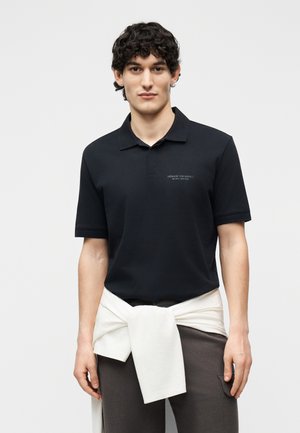 Giovane uomo con capelli ricci indossa una polo nera Armani Exchange e un maglione bianco legato in vita su sfondo semplice.