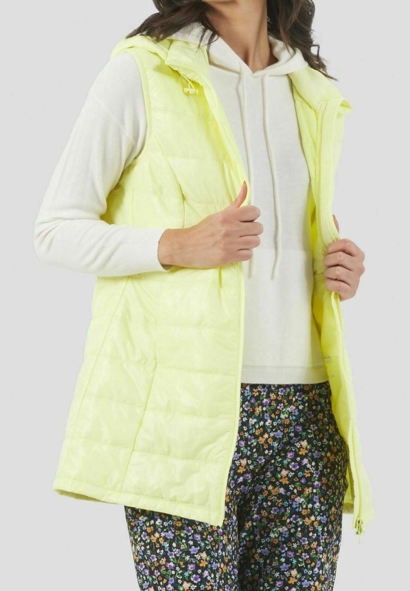 Femme portant un gilet matelassé sans manches jaune clair sur un sweat à capuche blanc et un pantalon à motifs floraux, ajustant la fermeture éclair du gilet.