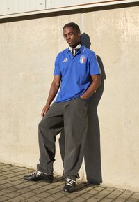 Μπλε πόλο μπλούζα με σήμα Ιταλίας, χρυσές λεπτομέρειες και κοντά μανίκια; συνδυασμένη με χαλαρές μαύρες τζιν και μαύρα αθλητικά παπούτσια με λευκές δέστρες.