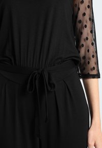 Black top featuring an elasticized waist with a tie. The sleeves are sheer fabric with polka dot pattern, contrasting with the solid body.