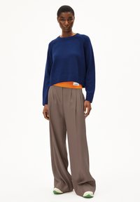 Navy knitted sweater with ribbed cuffs and hem, layered over an orange top. Loose beige trousers with wide legs and green-accented sneakers.
