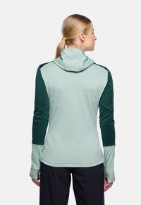 Frau mit blonden Haaren, die zu einem Pferdeschwanz gebunden sind, trägt einen langärmligen, hellgrünen und dunkelgrünen Kapuzenpullover, von hinten betrachtet.