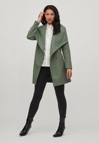 Abrigo verde de mezcla de lana con un frente largo y abierto, combinado con una camisa blanca de botones, jeans skinny negros y botas de tobillo negras puntiagudas.