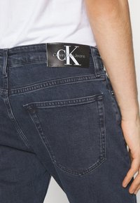 Mörk jeans med en åtsittande design, som har en svart lädretikett med "Calvin Klein Jeans"-logotypen vid midjan och en enkel bakficka.