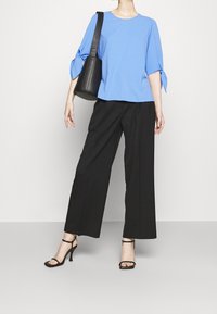 Maglietta blu a maniche corte con dettagli a fiocco, abbinata a pantaloni neri a gamba larga e sandali neri con tacco a fascia, mentre tiene una borsa nera.