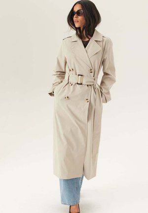 Vrouw draagt een beige trenchcoat met riem over blauwe spijkerbroek en zwarte schoenen, poseert met handen in de zakken, draagt donkere zonnebril.