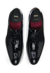 Een paar zwarte glanzende leren veterschoenen met bordeauxrode binnenzolen, gemarkeerd met "BOSS," van bovenaf weergegeven op een witte achtergrond.