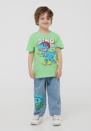 T-shirt verde in cotone con grafica di dinosauro; jeans blu a vestibilità rilassata con un design vivace di faccina sorridente; sneakers bianche.