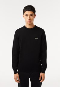 Maglione nero a maglia con maniche lunghe, polsini e orlo a coste, con un piccolo logo verde sul petto. Tessuto liscio e vestibilità classica.
