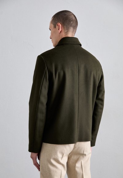 Olive green wool-blend jacket with a structured collar, tailored fit, and no visible fastenings, paired with beige trousers.