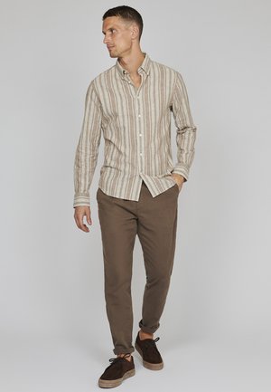 Man som bär en beige och vit randig skjorta med knappar, bruna byxor och mörkbruna skor, står och tittar åt sidan.