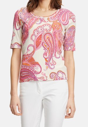 Mujer con una camiseta ajustada de manga corta con estampados de cachemira en tonos rosa y naranja, y pantalones blancos, de pie frente a un fondo liso.