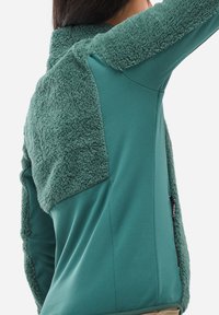 Eine Frau trägt eine türkisfarbene Jacke mit weichem, flauschigem Stoff auf dem oberen Rücken und den Ärmeln sowie glattem Stoff am unteren Rücken und an den Seiten.