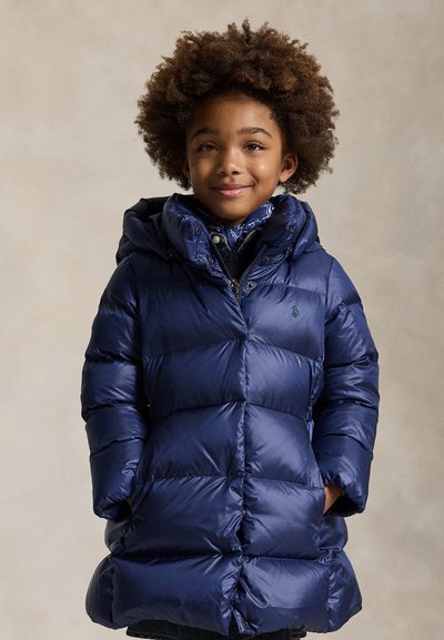 Bambino con capelli ricci che indossa una giacca imbottita blu e una sciarpa con motivo, sorridente con le mani nelle tasche, in piedi davanti a uno sfondo uniforme.