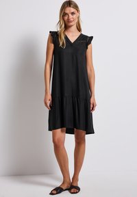 Schwarzes kurzes Kleid mit kurzen Ärmeln, V-Ausschnitt, Rüschenschultern und einem gestuften Saum. Glattes Material mit lockerem Schnitt, das bis zur Mitte des Oberschenkels reicht.