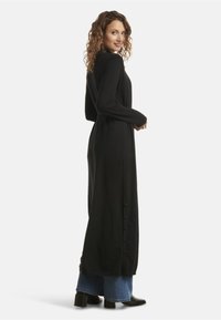 Woman with curly hair wearing a long black buttoned cardigan over blue jeans and black heeled boots, looking over her shoulder.