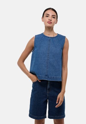 Jeune femme aux cheveux foncés portant un haut en denim sans manches et un short en denim foncé à hauteur de genoux, debout avec une main dans la poche.