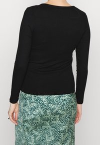 Person som bär en åtsittande svart långärmad topp och en högmidjad grön kjol med ett vitt paisleymönster, sedd bakifrån.