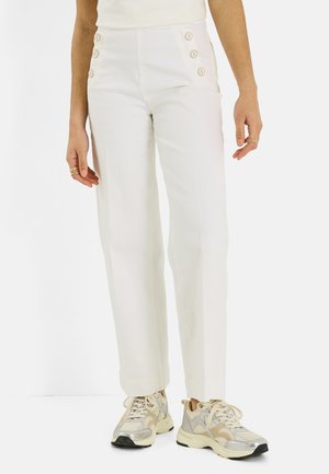 Femme portant un pantalon blanc taille haute avec des boutons sur les hanches et des baskets beige-blanc, debout devant un fond uni.