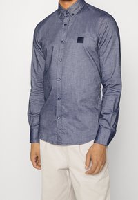 Navy blue button-up shirt in chambray fabric, featuring a small square pocket, long sleeves, and contrasting blue buttons.