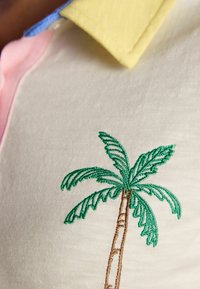 Nahaufnahme eines Poloshirts mit gelbem Kragen und grüner Palmenstickerei auf beigem Stoff.