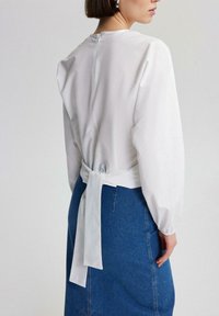 Frau in einer weißen Bluse mit langen Ärmeln und einem Rückenbindedetail, kombiniert mit einem blauen Jeansrock, seitlich und von hinten gezeigt.