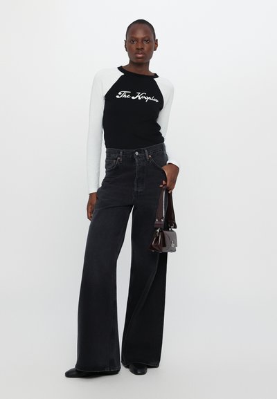 Black and white long-sleeve shirt with text, high-waisted black jeans, and small dark handbag; model stands with a neutral background.
