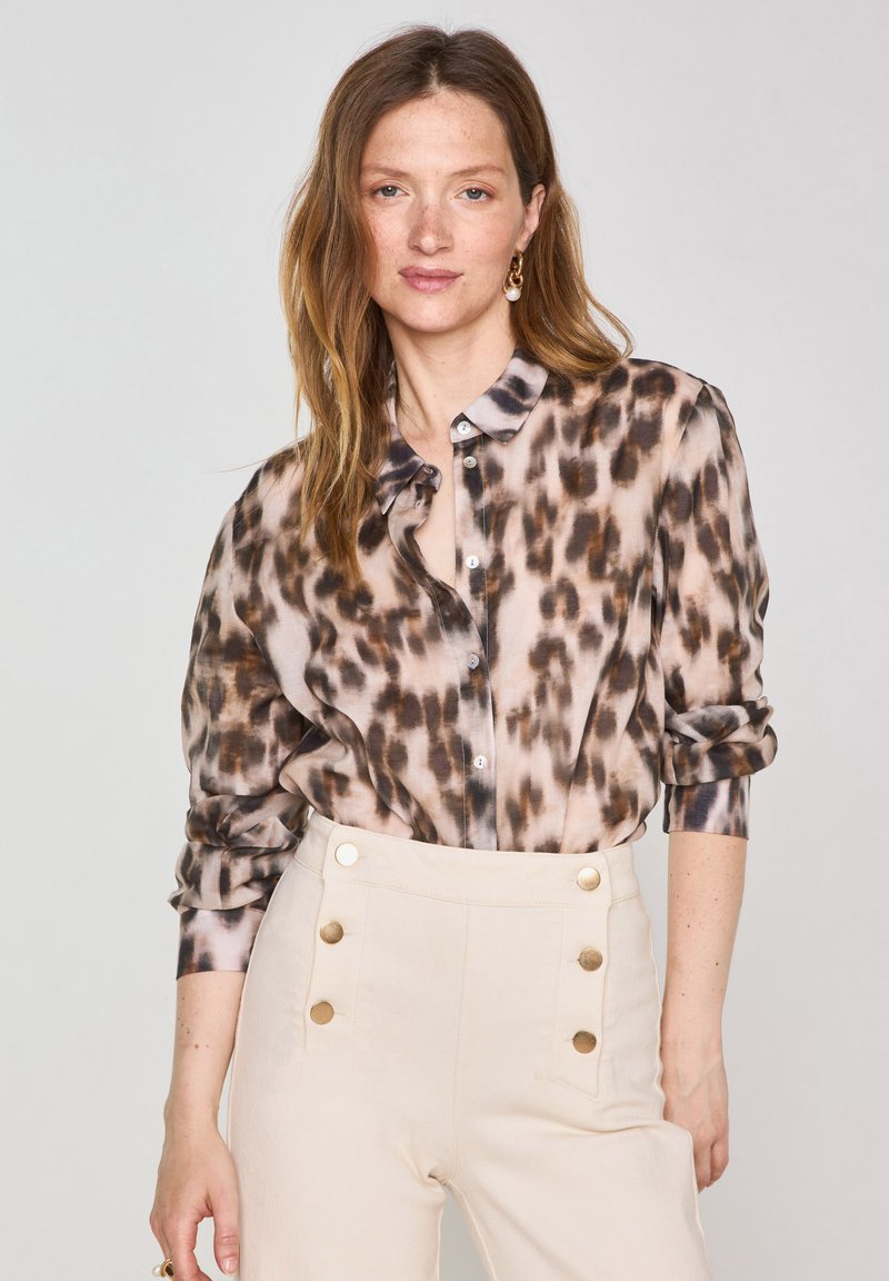 Camicia leggera e trasparente marrone e beige con motivo a leopardato, abbinata a pantaloni a vita alta color crema con bottoni dorati.