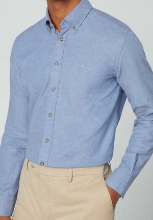 Homme portant une chemise bleu clair boutonnée avec col et pantalon beige, main dans la poche, montré du cou aux hanches.