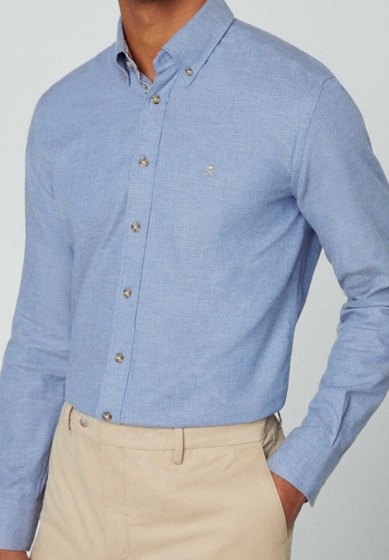 Homme portant une chemise bleu clair boutonnée avec col et pantalon beige, main dans la poche, montré du cou aux hanches.