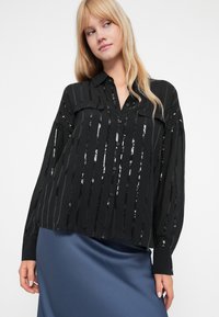 Blusa preta com riscas verticais de lantejoulas, fecho com botões na frente e mangas longas. Apresenta dois bolsos frontais e um corte descontraído.