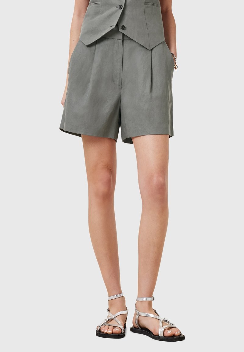 Personne portant un short gris taille haute et des sandales plates argentées à brides, debout les mains dans les poches devant un fond uni.