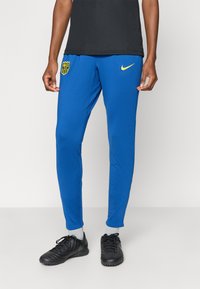 Μπλε αθλητικό παντελόνι από ελαστικό ύφασμα, με κίτρινο λογότυπο Nike και σήμα συλλόγου στον αριστερό μηρό. Στενή εφαρμογή, απαλό υλικό.