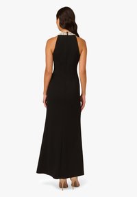 Schwarzes ärmelloses Kleid mit einer figurbetonten Silhouette und einem mit Perlen verzierten Kragen. Das Kleid endet bodenlang mit einer schlichten Form und einer Rückennaht.