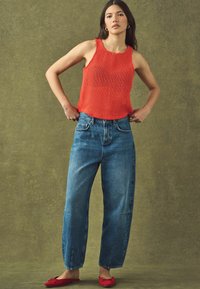 Rød strikket ærmeløs top, lyseblå højtaljede denimjeans med brede ben og røde flade sko mod en dæmpet grøn baggrund.
