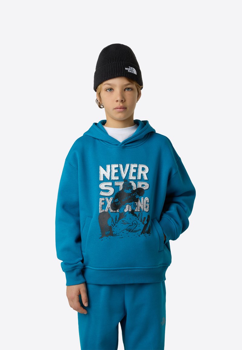 Modra jopa s kapuco z grafičnim tiskom z napisom "NEVER STOP EXPLORING," ki ima kengurujji žep in rebraste manšete. Skupaj z ujemajočimi modrimi hlačami.