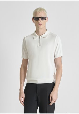 Hombre rubio con gafas de sol rectangulares negras, camisa blanca de manga corta con cuello y cremallera, y pantalones negros, de pie frente a un fondo liso.
