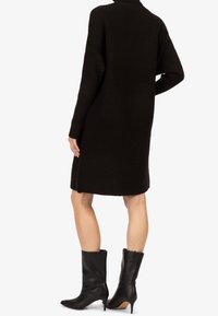 Robe noire en tricot côtelé à manches longues, longueur mi-cuisse, avec fentes latérales. Portée avec des bottes noires à talons aiguilles jusqu'au genou.