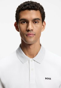 Camiseta polo blanca con un cuello de patrón hexagonal gris claro, tres botones y un logo negro "BOSS" en el lado izquierdo del pecho.