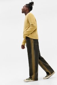 Ung person med dreadlocks iført en gul fluffy sweater, brede mørke jeans og grønne sneakers, som går sidelæns mod en hvid baggrund.
