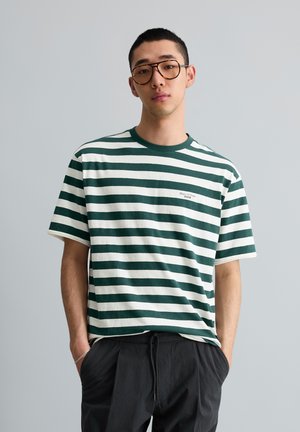 Junger Mann mit brauner Brille und grün-weiß gestreiftem T-Shirt mit Händen in den Taschen schwarzer Hosen, vor schlichtem hellgrauem Hintergrund.