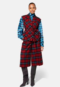 Rood en blauw geruite vest met een ceintuur in de taille, gedragen over een blauw geruit overhemd en rode geruite culottes, gecombineerd met grijze kniehoge laarzen.