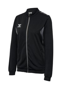 Schwarze Zip-Jacke mit hohem Kragen, kombiniert mit grauen seitlichen Akzenten und einem Logo auf der linken Brust. Strukturierter Stoff mit gerippten Bündchen.