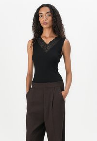 Top nero a coste senza maniche con scollatura a V in pizzo scallop. Abbinato a pantaloni alla moda marroni a vita alta con pieghe. Texture liscia.